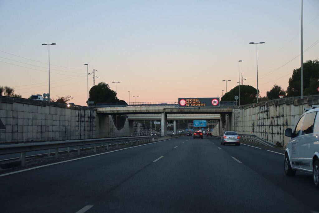 Cars driving on the A-7 motorway near Marbella at dusk, with an overhead DGT sign reading 'Mantenga distancia de seguridad' and an 80 km/h limit