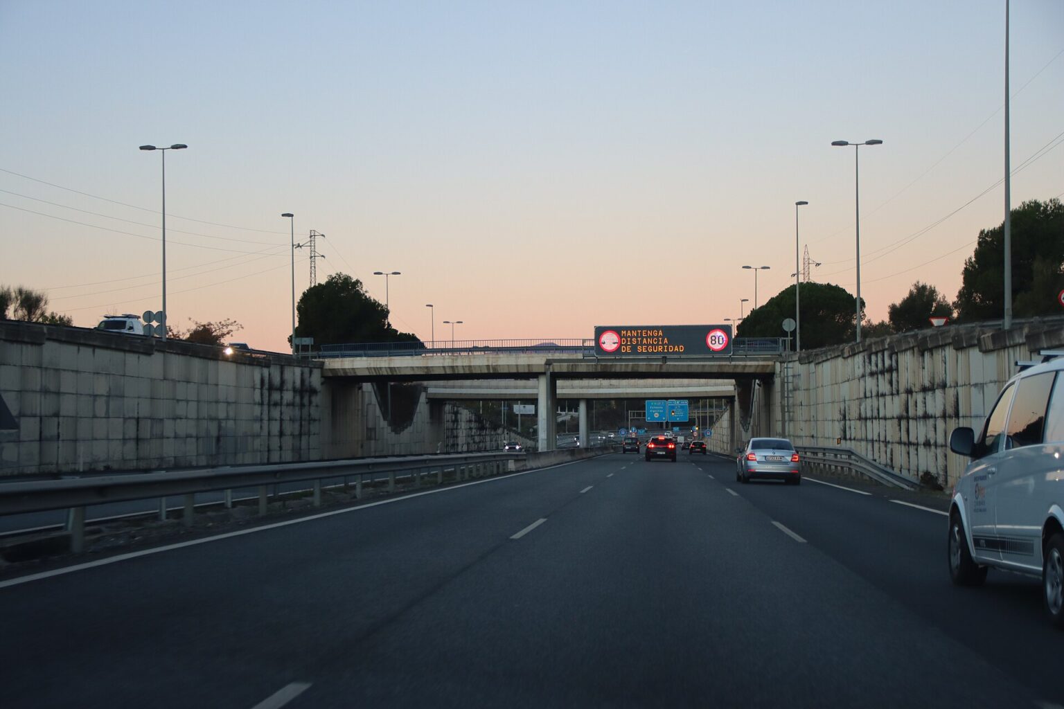 Cars driving on the A-7 motorway near Marbella at dusk, with an overhead DGT sign reading 'Mantenga distancia de seguridad' and an 80 km/h limit